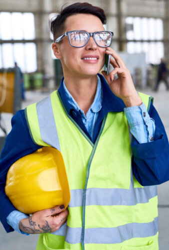 Portrait of modern female worker speaking by phone and holding hardhat in workshop of industrial plant