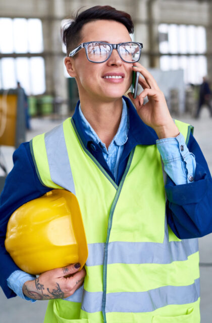 Portrait of modern female worker speaking by phone and holding hardhat in workshop of industrial plant